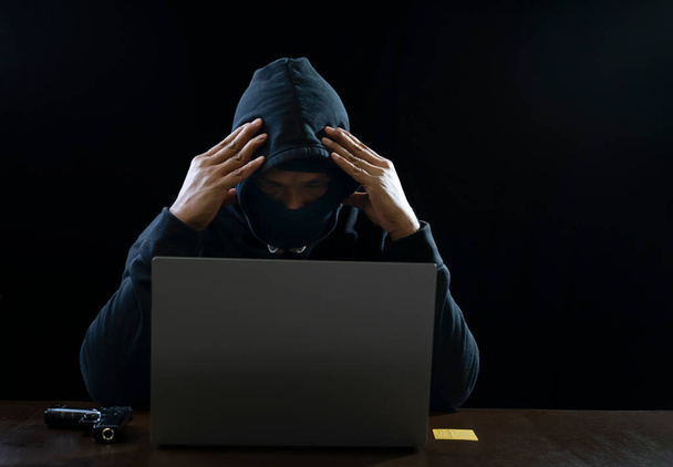 a stress hacker in black hoodie doing both hands on head is hacking failed,there are device such as gun,credit card and laptop on table isolated on dark background - Foto, Imagem