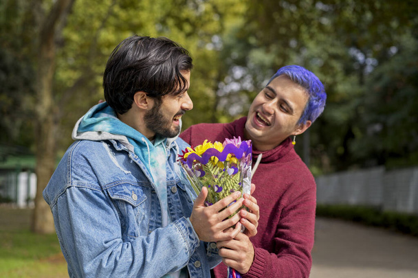 Пара латиноамериканських геїв у парку обіймають один одного. Один дивує іншого букетом квітів. - Фото, зображення