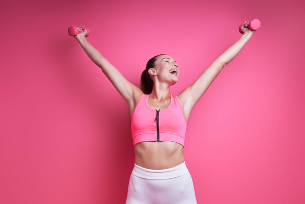 Happy young woman in sports clothing exercising with dumbbells against pink background - Foto, afbeelding