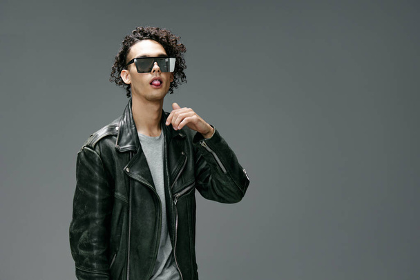 Side of view of stylish tanned curly man leather jacket trendy sunglasses posing isolated on gray studio background. Cool fashion offer. Huge Seasonal Sale New Collection concept. Copy space for ad - Photo, Image
