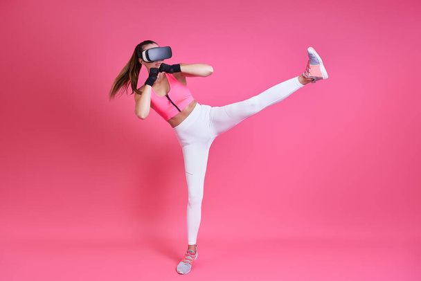 Young woman in virtual reality glasses kicking against pink background - Valokuva, kuva