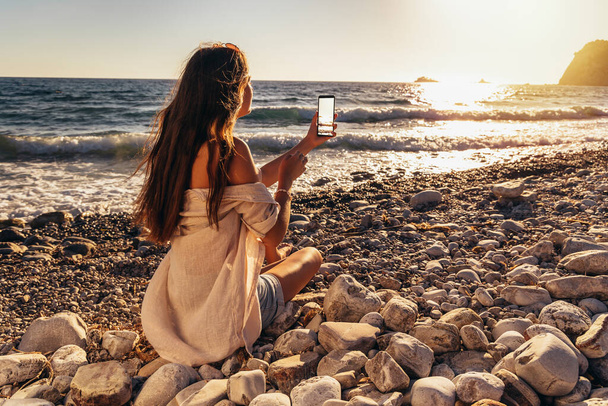 Teenage girl pictures on the phone a beautiful view of the sea and the sunset. Concept of travel, vacation - 写真・画像