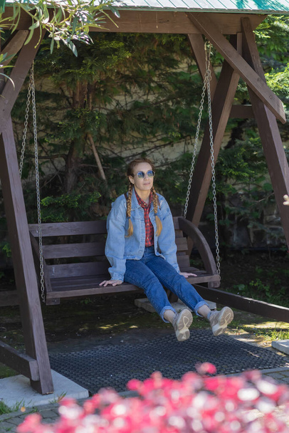 Жінка з кісками в джинсовому вбранні гойдається з ногою вгору на дерев'яних гойдалках в тінистій зоні парку - Фото, зображення