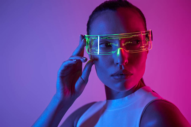 Beautiful young woman adjusting futuristic glasses against dark background - Photo, image