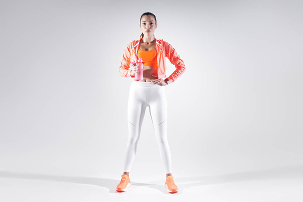 Full length of beautiful young woman in sports clothing drinking water against white background - Фото, зображення