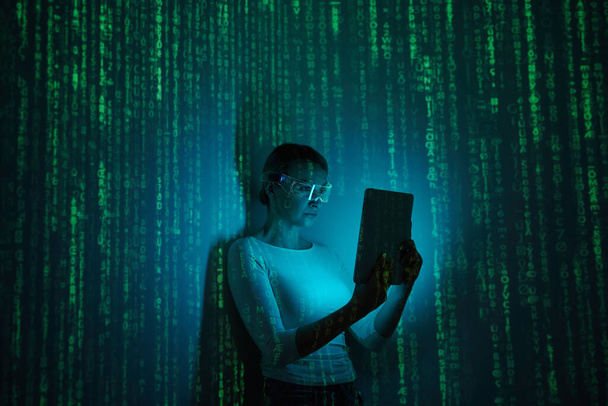 Confident woman in futuristic glasses using digital tablet against dark background with green symbols - Фото, изображение
