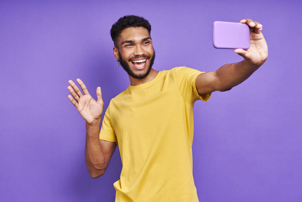 Playful African man looking at smart phone and waving hand against purple background - Photo, Image