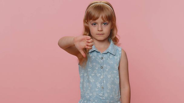 Dislike. Upset young preteen child girl kid showing thumbs down sign gesture, expressing discontent, disapproval, dissatisfied bad work indoors. Displeased toddler children on pink studio background - Photo, Image
