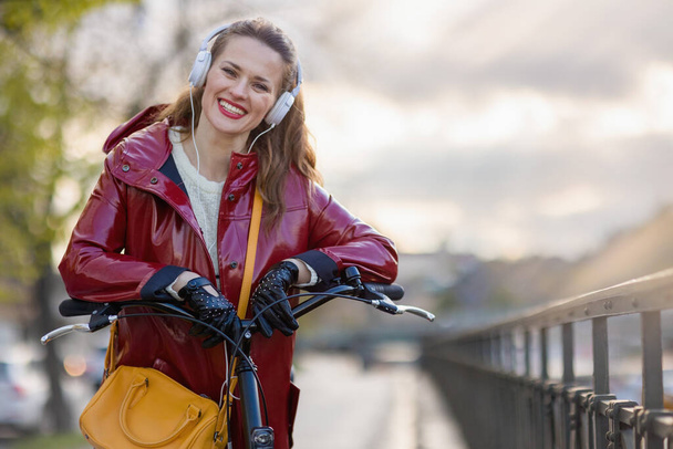 Portrait of smiling stylish female in red rain coat with bicycle listening to the music with headphones outside on the city street. - Zdjęcie, obraz