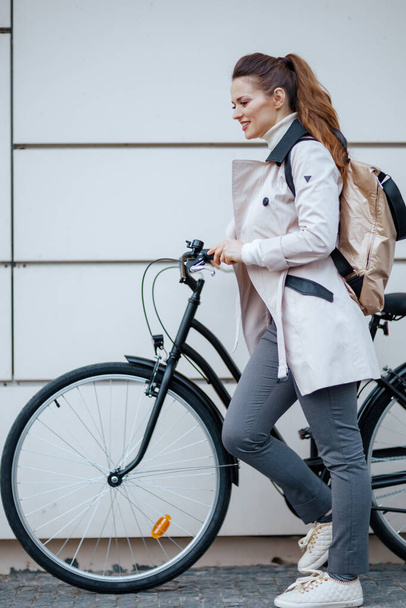 Full length portrait of happy elegant middle aged woman in beige trench coat with bicycle and backpack In front of a metal wall outside on the city street. - Zdjęcie, obraz
