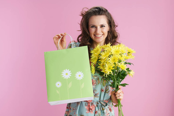 Portrait of happy stylish female in floral dress with yellow chrysanthemums flowers and green shopping bag isolated on pink background. - Foto, Imagem