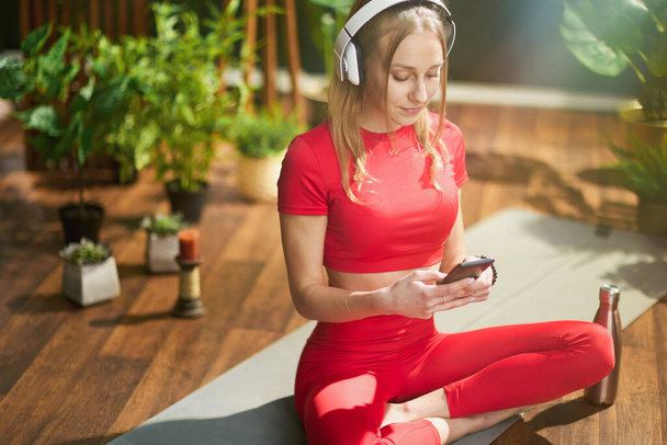 young female in red fitness clothes with yoga mat and smartphone listening to the music with headphones in the modern green house. - Фото, изображение