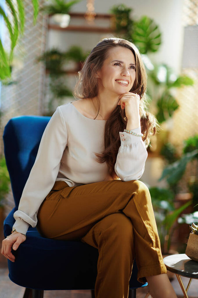 Green Home. happy trendy middle aged housewife with long wavy hair in the modern house in sunny day in green pants and grey blouse sitting in a blue armchair. - Photo, image