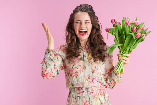 cheerful 40 years old woman in floral dress with tulips bouquet isolated on pink background. - Photo, Image