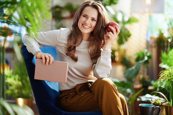Green Home. smiling young woman with long wavy hair with book and red apple in green pants and grey blouse sitting in a blue armchair in the modern house in sunny day. - Fotografie, Obrázek