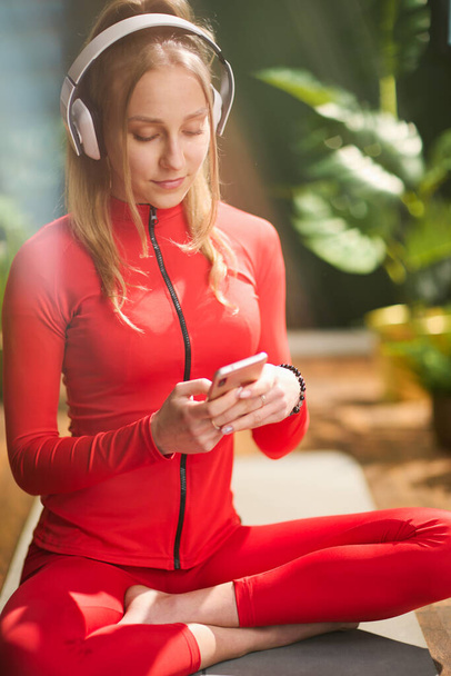 modern female in red fitness clothes with smartphone listening to the music with headphones in the modern green living room. - Fotografie, Obrázek