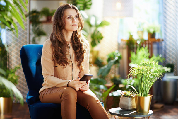Green Home. pensive middle aged woman with long wavy hair in the modern house in sunny day in beige pants and blouse using smartphone applications while sitting in a blue armchair. - Photo, image
