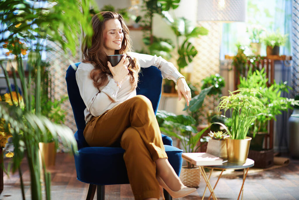 Groen Huis. lachende 40-jarige vrouw met lang golvend haar met kopje warme chocolademelk in groene broek en grijze blouse zittend in een blauwe fauteuil in het moderne huis op zonnige dag. - Foto, afbeelding
