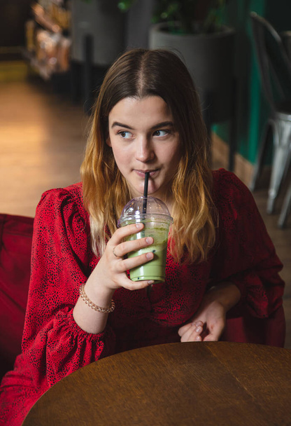 Молодая женщина в красном платье в кафе пьет летний зеленый напиток латте со льдом. - Фото, изображение