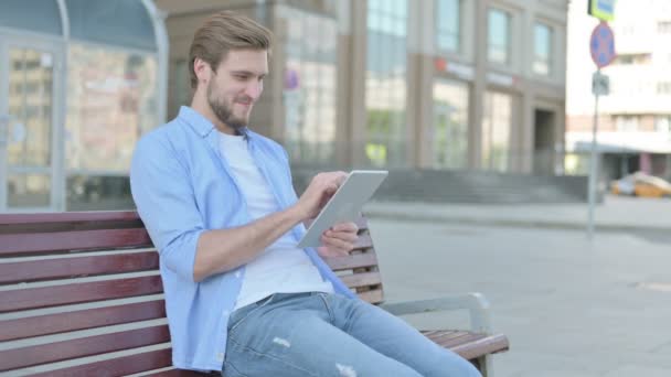 Hombre de mediana edad celebrando victoria en línea en la tableta mientras está sentado al aire libre en el banco - Metraje, vídeo