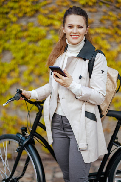smiling elegant woman in beige trench coat with bicycle and backpack using smartphone app against the green wall outside in the city. - Foto, afbeelding