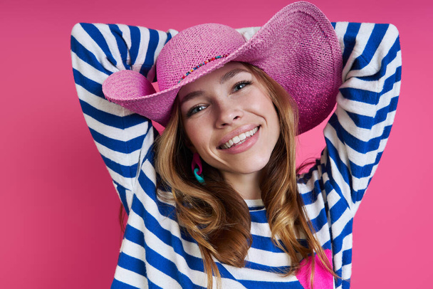 Beautiful woman in pink hat holding hands behind head while standing against colored background - Foto, imagen
