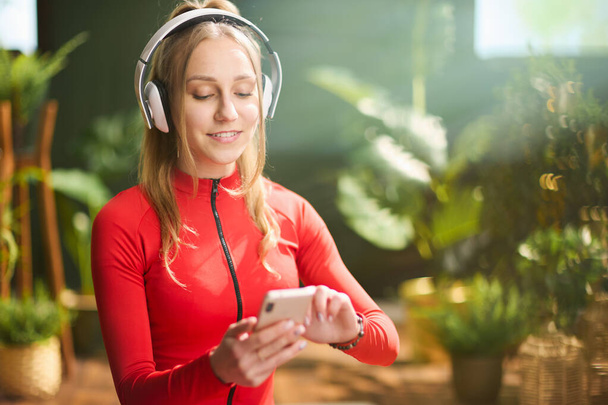 smiling young female in red fitness clothes with headphones using smartphone at modern green home. - Φωτογραφία, εικόνα