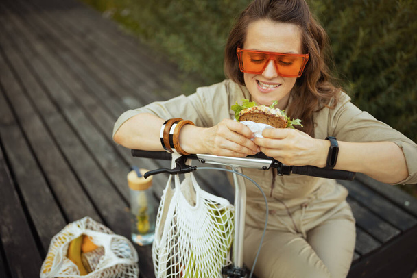 smiling elegant female in sunglasses and overall with string bag, scooter and sandwich sitting outdoors in the city. - Foto, Imagen