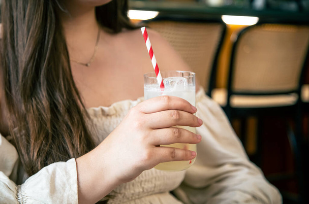 Een glas koude limonade met een gestreept rietje in een vrouwenhand in een café, het concept van zomerdrankjes. - Foto, afbeelding