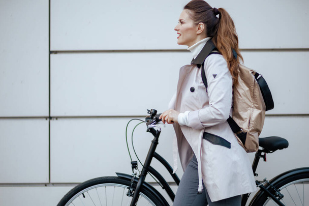 elegant female in beige trench coat with bicycle and backpack In front of a metal wall outdoors in the city. - Foto, imagen