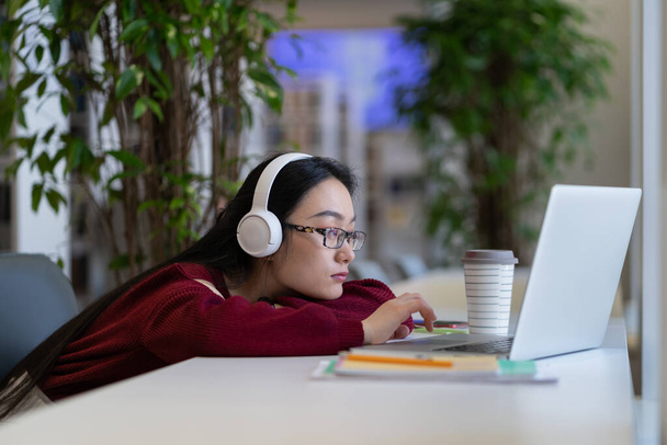 Unavená znuděná asijská studentka ve sluchátkách surfující na internetu na notebooku při studiu v moderní univerzitní knihovně, selektivní zaměření. Procrastination v online učení, distanční vzdělávání - Fotografie, Obrázek