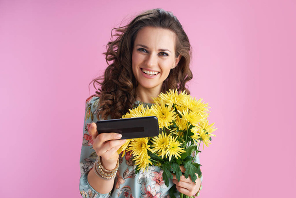 Portrait of smiling modern 40 years old woman in floral dress with yellow chrysanthemums flowers sending text message using smartphone isolated on pink background. - Photo, image