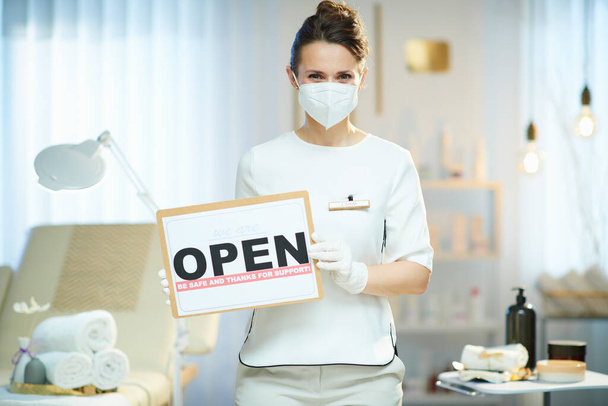 Business during covid-19 pandemic. female employee with ffp2 mask and open sign in modern beauty studio. - 写真・画像