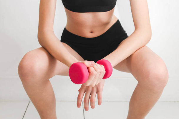 Young beautiful caucasian girl in fitness holds one hand pink one kilogram dumbbell and crouches against the background of a white wall, close-up side view.Concept of home fitness and healthy lifestyle. - Fotoğraf, Görsel