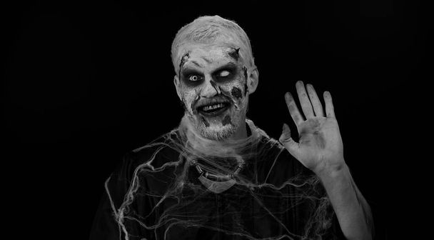 Жуткий человек с окровавленным лицом, макияж зомби на Хэллоуин. Страшный раненый нежить парень улыбается дружелюбно перед камерой и машет руками жестом приветствия или прощания, приветствуя с гостеприимным выражением - Фото, изображение