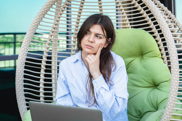 Σοβαρή νεαρή γυναίκα εργάζεται σε ένα φορητό υπολογιστή, ενώ κάθεται σε μια αιώρα στη βεράντα του γραφείου. - Φωτογραφία, εικόνα