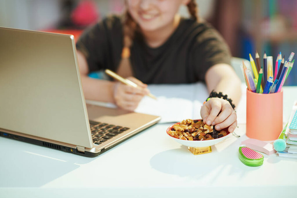 Closeup on smiling girl in grey shirt with laptop homeschooling and eating healthy snack at home in sunny day. - Foto, imagen