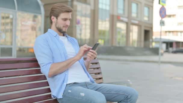 Középkorú férfi ünnepli Online siker Smartphone, miközben ül a szabadban a padon - Felvétel, videó