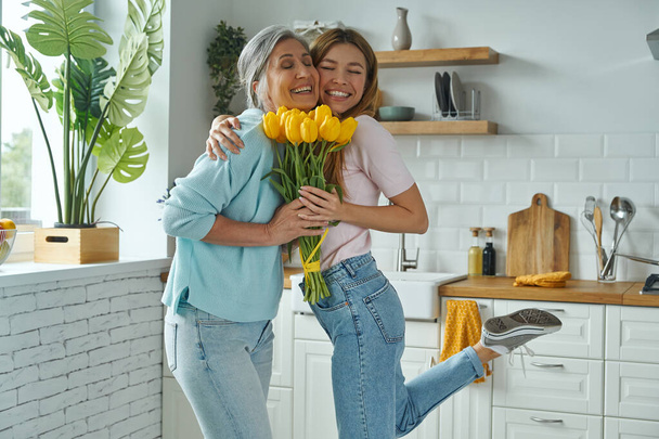 Happy senior mother receiving a bunch of tulips from her daughter while standing at the kitchen - Fotoğraf, Görsel