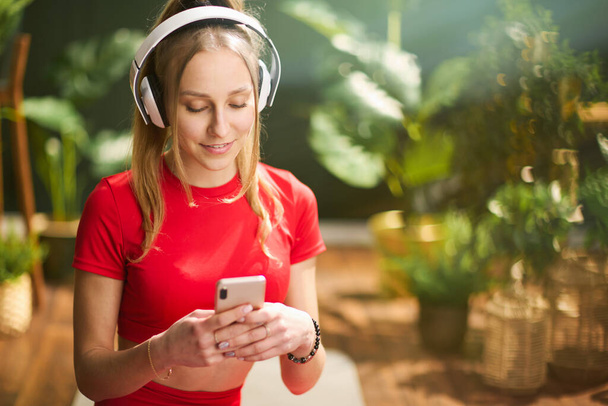 smiling young female in red fitness clothes with smartphone and headphones in the modern green house. - Φωτογραφία, εικόνα