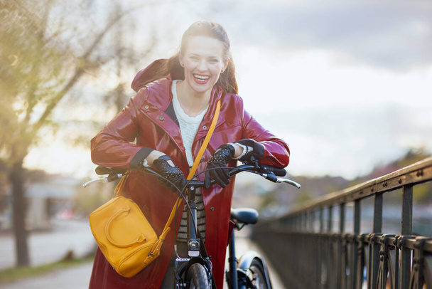 Portrait of smiling elegant 40 years old woman in red rain coat with bicycle outside in the city. - Foto, imagen