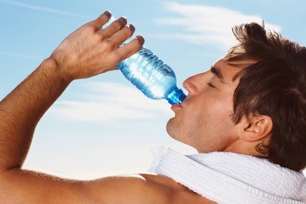 Nahaufnahme eines gutaussehenden Typen, der nach einem Training Wasser mit einem Handtuch um den Hals trinkt. - Foto, Bild