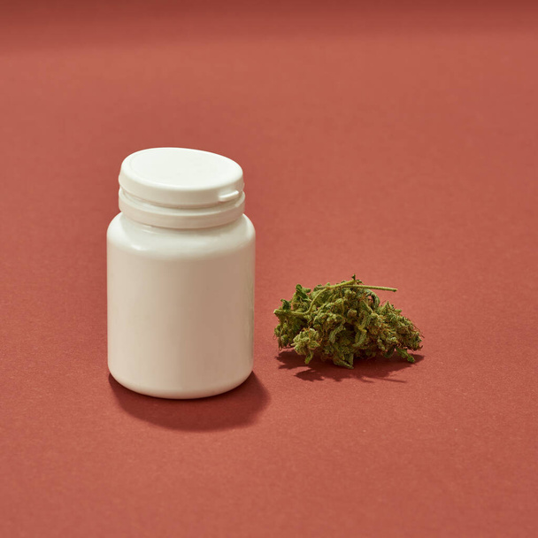Fresh green dry cutting marijuana weed and plastic jar isolated on red background. Light drug and addiction. Herbal medicine and painkiller therapy. Natural organic cannabis. Copy space - Photo, image
