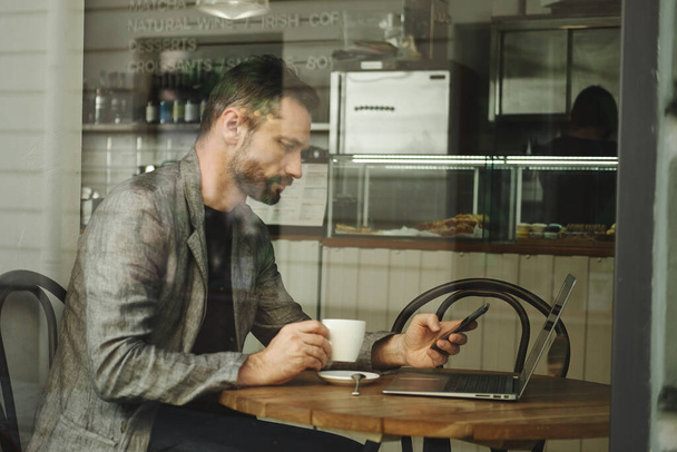 Caucasian man faceless working on a laptop behind the glass in a cafe. The man drinks coffee and looks at the phone. Multi-screening concept - 写真・画像