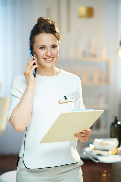 smiling middle aged woman worker with clipboard speaking on a smartphone in modern beauty studio. - Foto, Imagen