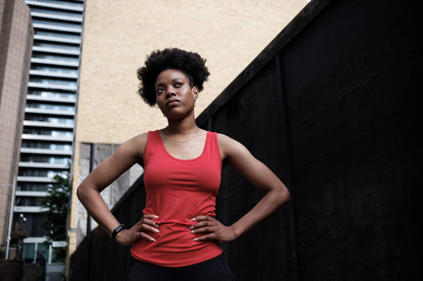 A young black woman is doing exercise outdoor in London. She is wearing red sleeveless t-shirt, black leggings and curly hair. - Fotografie, Obrázek
