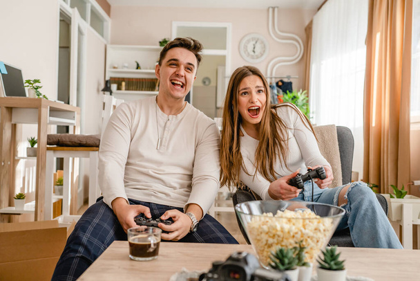дві кавказькі люди, сидячи вдома, грають в ігрову консоль, веселяться молода пара чоловік і жінка хлопець і дівчина проводять час разом, тримаючи джойстик контролер усміхнений вид спереду
 - Фото, зображення