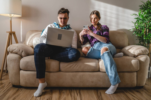 Two people man and woman young adult caucasian couple boyfriend and girlfriend or married husband and wife sitting on the sofa bed at home making a phone call or online purchase internet on laptop - Foto, Imagem