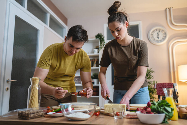 Two people young adult couple man and woman husband and wife or boyfriend and girlfriend preparing pizza food at home helping each other cutting and add ingredients and spices real people copy space - Foto, Imagen