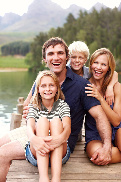 Smiling and laughing family sitting together on a jetty at a lake. - 写真・画像